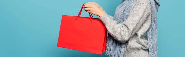 Vue recadrée de la femme portant écharpe et pull, tenant des sacs à provisions rouges isolés sur bleu, bannière — Photo de stock