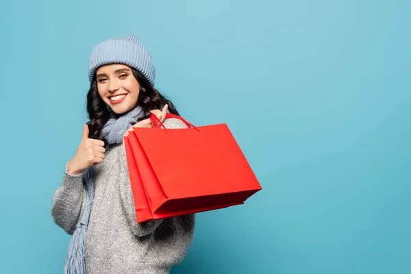 Feliz jovem mulher adulta com sacos de compras vermelhas mostrando polegar para cima enquanto olha para a câmera isolada no azul — Fotografia de Stock