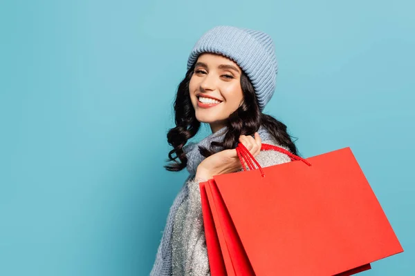 Sonriente mujer adulta joven en sombrero y bufanda sosteniendo bolsas de compras rojas y mirando a la cámara aislada en azul - foto de stock