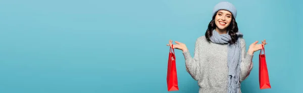 Mulher morena sorridente de chapéu e cachecol olhando para a câmera enquanto segurava sacos de compras vermelhos isolados em azul, banner — Fotografia de Stock