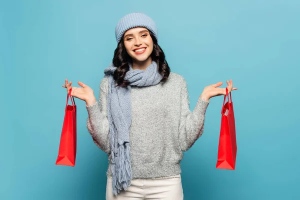 Vista frontal da mulher sorridente em roupa de inverno olhando para a câmera enquanto segurando sacos de compras vermelhos isolados no azul — Fotografia de Stock