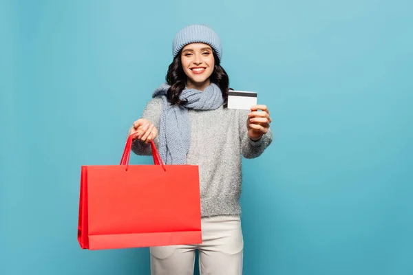Vista frontal da mulher feliz em roupa de inverno olhando para a câmera enquanto mostra cartão de crédito e sacos de compras vermelhos isolados no azul — Fotografia de Stock