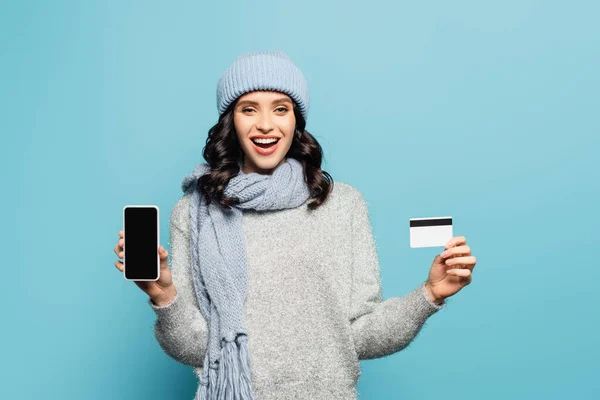Захоплена брюнетка в зимовому вбранні показує смартфон і кредитну картку, дивлячись на камеру ізольовано на синьому — стокове фото