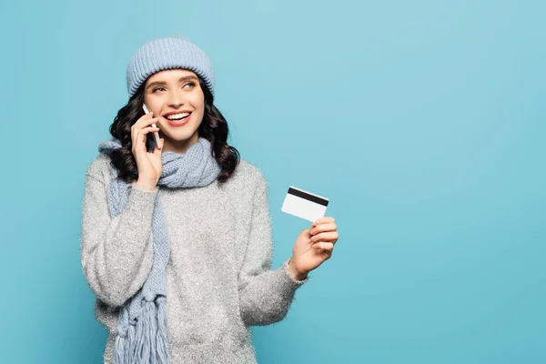 Mujer morena feliz en bufanda y sombrero hablando en el teléfono inteligente mientras mantiene la tarjeta de crédito aislada en azul - foto de stock