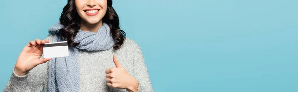 Vista recortada de la mujer morena con bufanda que muestra el pulgar hacia arriba mientras sostiene la tarjeta de crédito aislada en azul, pancarta - foto de stock