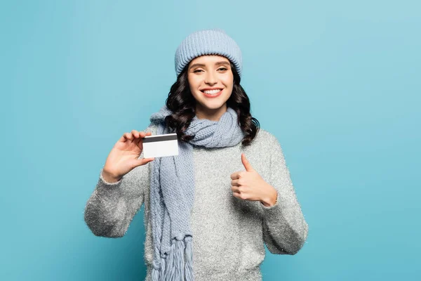 Mujer morena feliz en traje de invierno con tarjeta de crédito que muestra el pulgar hacia arriba mientras mira la cámara aislada en azul - foto de stock