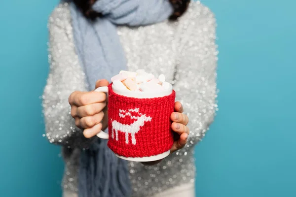 Close up view of cup with marshmallows and knitted holder in hands of blurred woman on background isolated on blue — Stock Photo