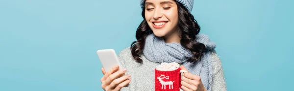 Cheerful brunette woman in scarf looking at smartphone while holding cup with knitted holder isolated on blue, banner — Stock Photo