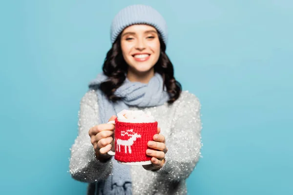 Cup with marshmallows and knitted holder in hands of blurred woman on background isolated on blue — Stock Photo