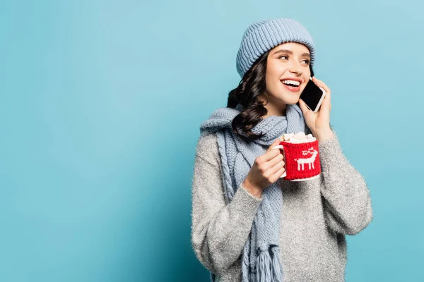 Happy woman in scarf and hat talking on smartphone while holding cup with marshmallows and knitted holder isolated on blue — Stock Photo