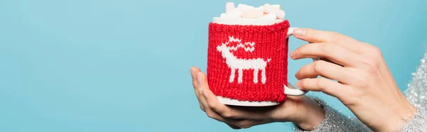 Close up view of cup with marshmallows and knitted holder in hands of woman isolated on blue, banner — Stock Photo