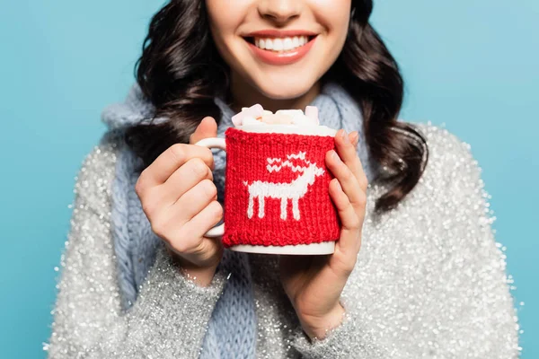 Vista recortada de mujer morena en bufanda sosteniendo taza con malvaviscos y titular de punto aislado en azul - foto de stock