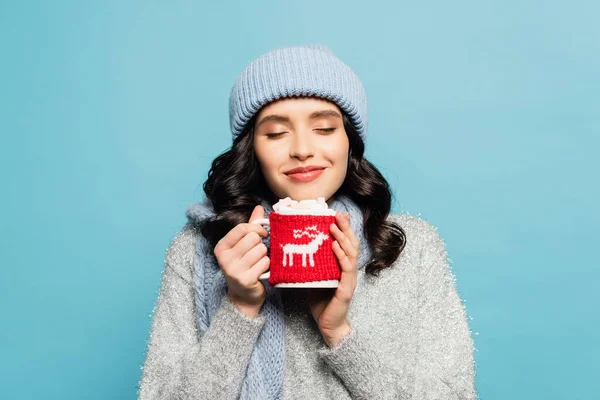 Donna bruna sorridente in abito invernale, con gli occhi chiusi in possesso di tazza con marshmallow isolati su blu — Foto stock