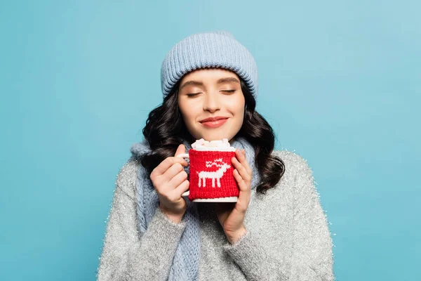 Mujer morena sonriente en traje de invierno, con los ojos cerrados sosteniendo taza con malvaviscos aislados en azul - foto de stock