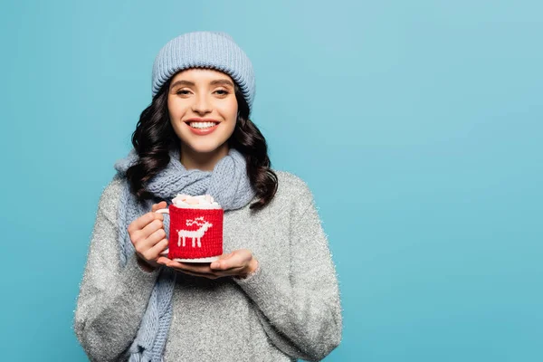 Femme heureuse en chapeau et écharpe regardant la caméra tout en tenant la tasse avec des guimauves isolées sur bleu — Photo de stock