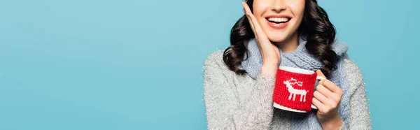 Cropped view of brunette woman with hand on cheek holding cup with knitted holder isolated on blue, banner — Stock Photo