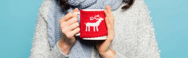 Cropped view of woman in scarf and pullover holding cup with knitted holder isolated on blue, banner — Stock Photo