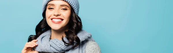 Portrait of cheerful brunette woman in hat and scarf looking at camera isolated on blue, banner — Stock Photo