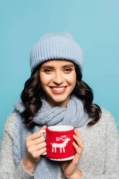 Heureuse femme brune en chapeau et écharpe regardant la caméra tout en tenant le capuchon avec support tricoté isolé sur bleu — Photo de stock