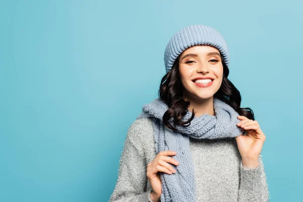 Glückliche brünette Frau im winterlichen Outfit, die Schal berührt, während sie isoliert auf blau in die Kamera schaut — Stockfoto