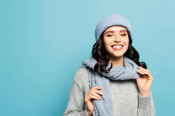 Heureuse femme brune en tenue d'hiver touchant écharpe tout en regardant la caméra isolée sur bleu — Photo de stock