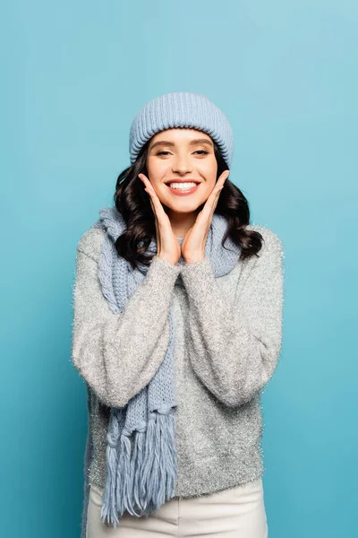 Femme heureuse avec les mains près des joues portant une tenue d'hiver et regardant la caméra isolée sur bleu — Photo de stock