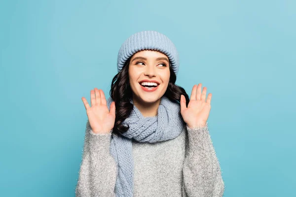 Mujer morena alegre en sombrero y bufanda mostrando las palmas mientras mira hacia otro lado aislado en azul - foto de stock