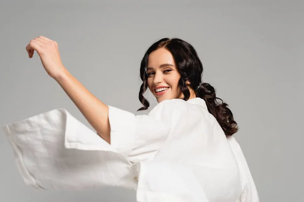 Femme brune souriante en chemise blanche regardant la caméra tout en se retournant isolé sur gris — Photo de stock