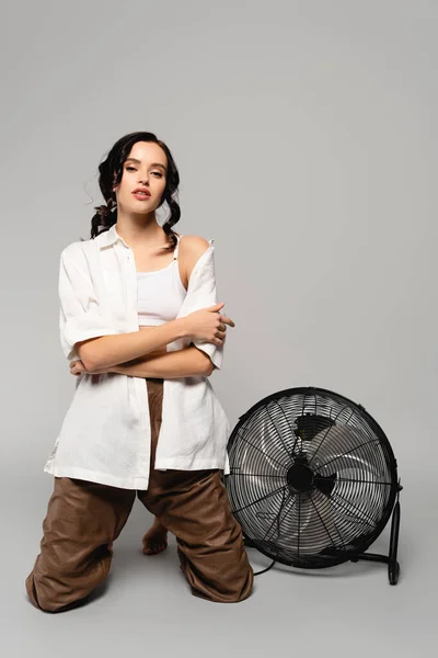 Full length of brunette woman with crossed arms looking at camera while standing on knees near fan on grey — Stock Photo