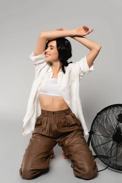 Femme brune souriante avec les mains dans l'air, debout sur les genoux près du ventilateur sur le gris — Photo de stock