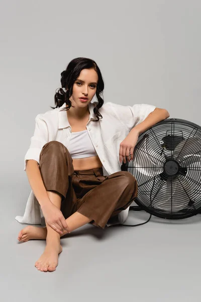Pleine longueur de femme brune en vêtements décontractés avec les jambes croisées s'appuyant sur le ventilateur sur gris — Photo de stock