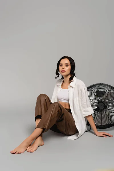 Full length of brunette woman with crossed legs looking at camera while sitting near fan on grey — Stock Photo