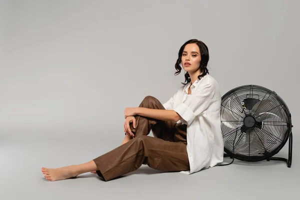 Comprimento total de mulher morena encaracolada em camisa e calças de couro olhando para a câmera e abraçando o joelho perto do ventilador no cinza — Fotografia de Stock