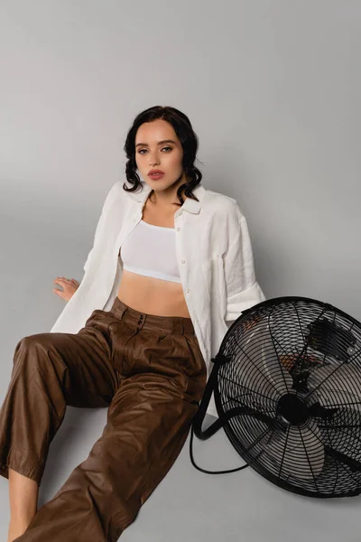 Brunette woman in shirt and leather pants looking at camera while sitting near fan on grey — Stock Photo