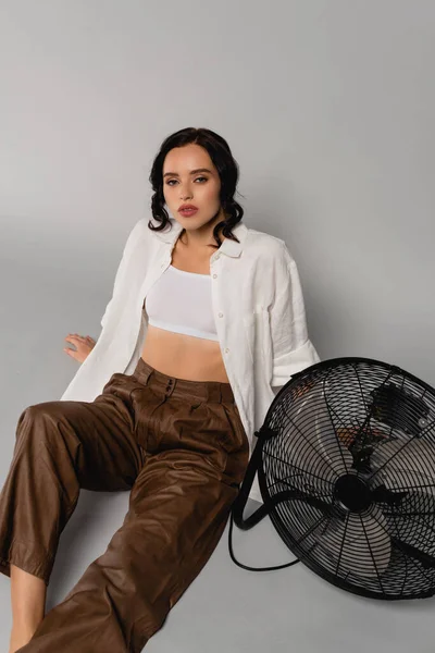 Brunette femme en chemise et pantalon en cuir regardant la caméra tout en étant assis près du ventilateur sur gris — Photo de stock
