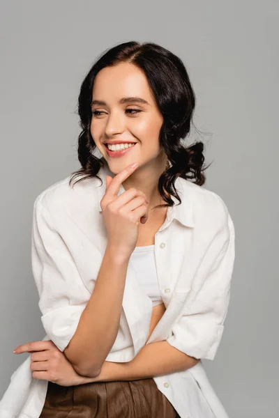 Mujer morena sonriente en top blanco y camisa mirando hacia otro lado aislado en gris - foto de stock