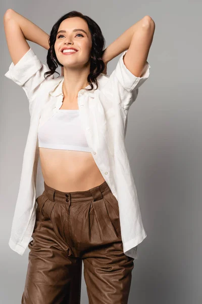 Mujer morena feliz con las manos detrás de la cabeza mirando hacia arriba aislado en gris - foto de stock