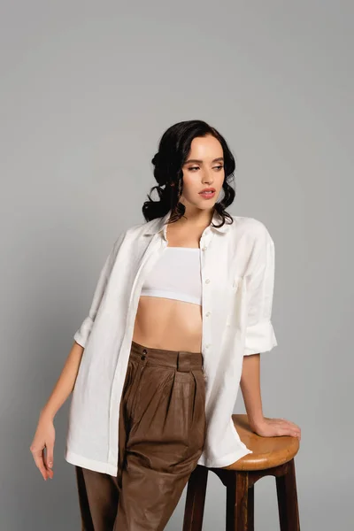 Brunette woman in shirt and pants looking away while leaning on stool isolated on grey — Stock Photo