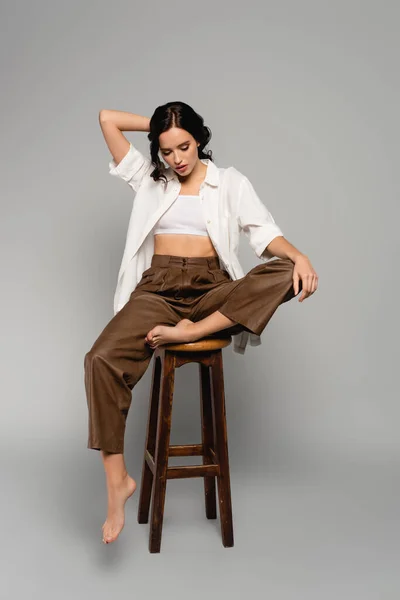 Full length of brunette woman in casual clothes posing while sitting on stool on grey — Stock Photo