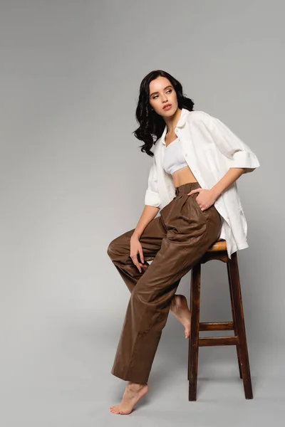 Femme brune pleine longueur avec la main dans la poche, regardant loin tout en posant sur le tabouret sur gris — Photo de stock