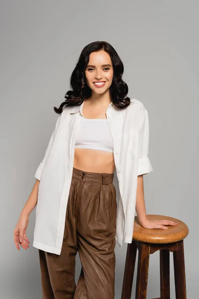 Smiling brunette woman in white top and shirt looking at camera while leaning on stool isolated on grey — Stock Photo