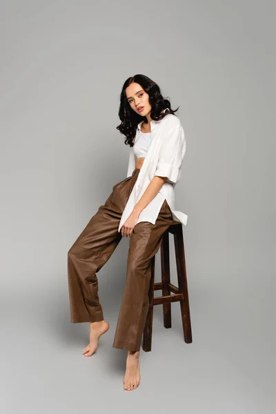 Longitud completa de la mujer morena rizada en camisa y pantalones de cuero posando mientras está sentado en el taburete sobre fondo gris - foto de stock