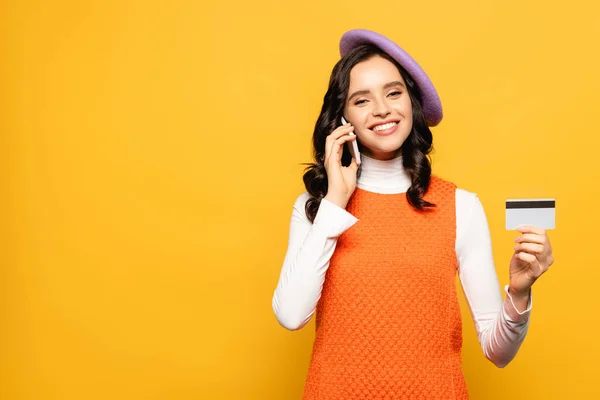 Mujer morena sonriente en boina hablando en smartphone y mirando a la cámara mientras muestra la tarjeta de crédito aislada en amarillo - foto de stock