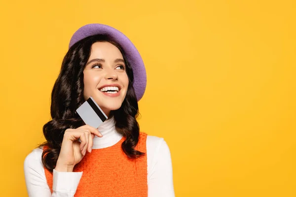 Cheerful brunette woman in beret looking up while holding credit card isolated on yellow — Stock Photo