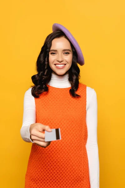 Front view of smiling brunette woman in beret looking at camera while giving credit card isolated on yellow on blurred foreground — Stock Photo