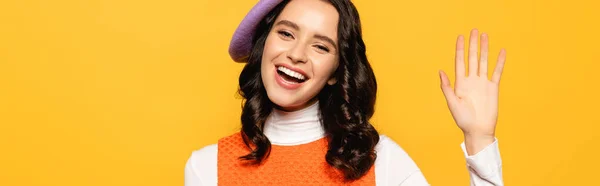 Happy brunette woman in beret with waving hand looking at camera isolated on yellow, banner — Stock Photo