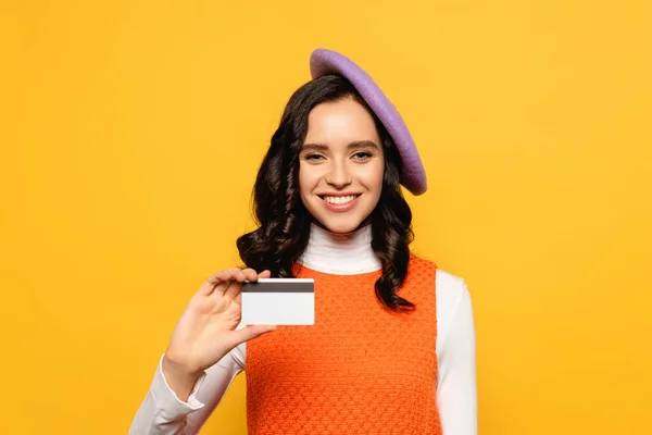 Mujer morena sonriente en boina mirando a la cámara mientras muestra la tarjeta de crédito aislada en amarillo - foto de stock