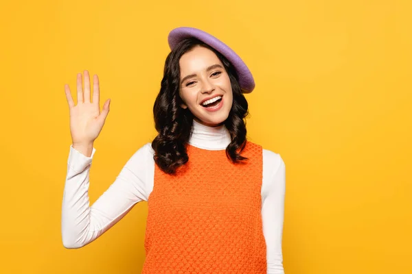 Cheerful brunette woman in beret with waving hand looking at camera isolated on yellow — Stock Photo