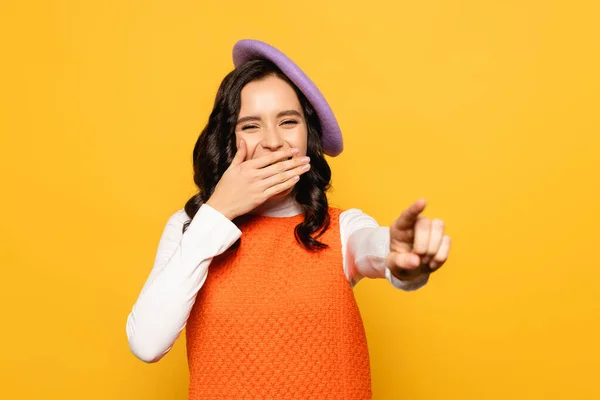 Brunette woman in beret covering mouth with hand and pointing with finger isolated on yellow on blurred foreground — Stock Photo