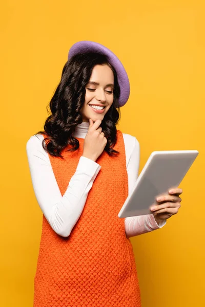 Happy brunette woman in beret thinking while looking at digital tablet isolated on yellow — Stock Photo